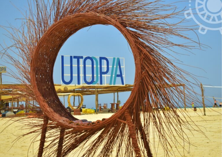 Супер Oстров Утопия Из Хургады 19 A straw arch frames the “UTOPIA” sign on bright white sand beneath a clear blue sky near Hurghada and Safaga.