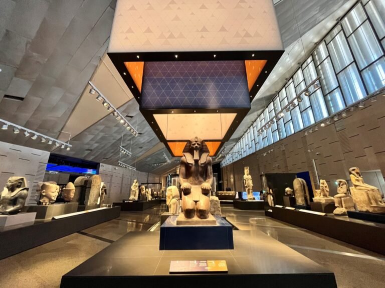 Каир автобусом из Хургады с посещением Grand Egyptian Museum (Новый музей) + Прогулка по Нилу 7 Inside the Grand Egyptian Museum, Ramses II statue stands in a modern gallery of gold-toned Egyptian sculptures under warm orange lights.