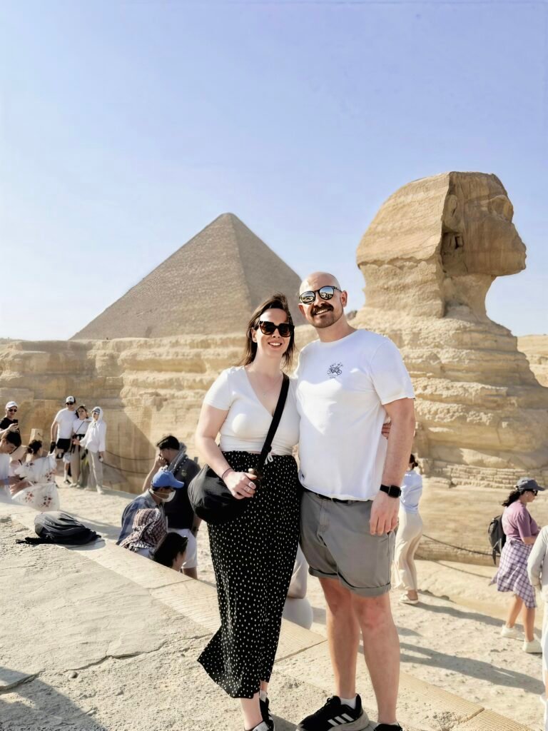 Каир на автобусе из Шарм Эль Шейха 2 Couple posing in front of the Giza pyramids and the Sphinx in bright daylight, wearing sunglasses and white clothing.
